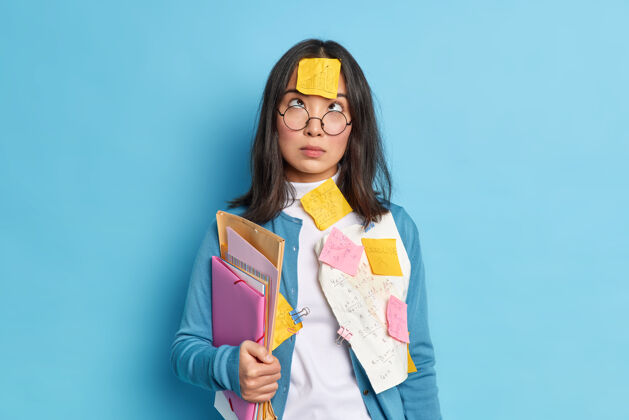 沮丧深色头发的亚洲女学生摄影棚用贴纸夹在额头上集中学习数学文件夹蓝色一个