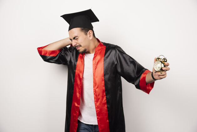 时钟年轻的研究生拿着钟在白地上感到痛苦帽子毕业男人