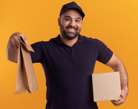 中年微笑着的中年送货员 穿着制服 戴着帽子 拿着盒子 拿着隔离在黄色墙上的纸食品包制服男人食物