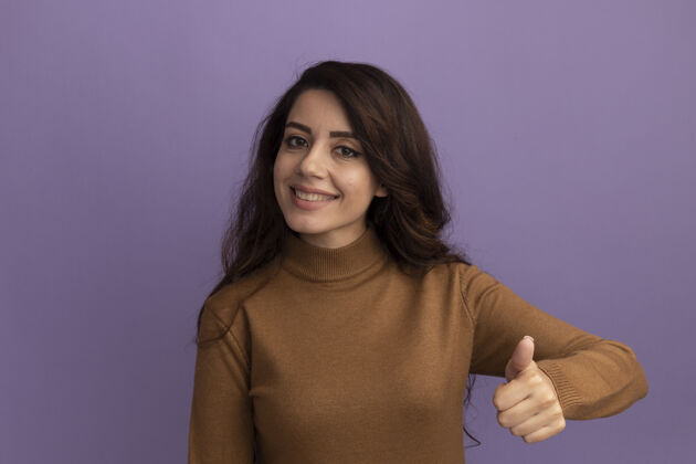高领毛衣微笑的年轻漂亮女孩穿着棕色高领毛衣 在紫色的墙上孤立地竖起大拇指秀年轻穿