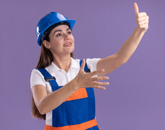 拇指微笑着看着身穿制服的年轻建筑妇女在紫色的墙上孤立地竖起大拇指女人微笑建筑工人