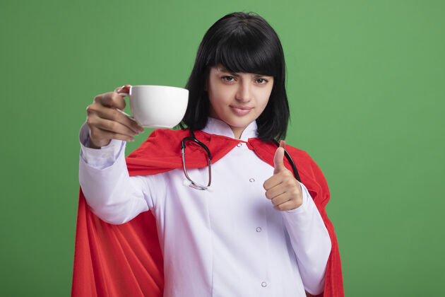 举行自信的年轻超级英雄女孩戴着听诊器 穿着医用长袍和斗篷 手里拿着一杯茶 在绿色的墙上孤立地竖起大拇指超级英雄斗篷茶