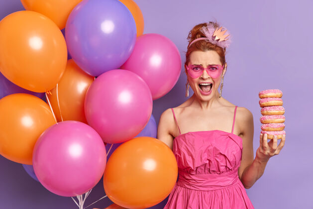奇迹愤怒的姜女 穿着粉色节日礼服 戴着心形太阳镜 手里拿着一堆甜甜圈 大声喊道 手里拿着五颜六色的氦气球喊年轻堆
