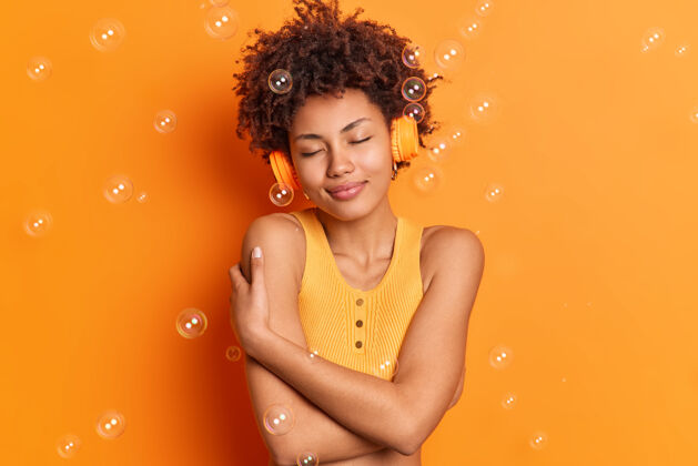 一个高兴的非裔美国妇女拥抱自己赤裸的肩膀站着满意地闭上眼睛通过立体声耳机听音乐摆在橙色的工作室墙壁与肥皂泡周围音乐请非洲