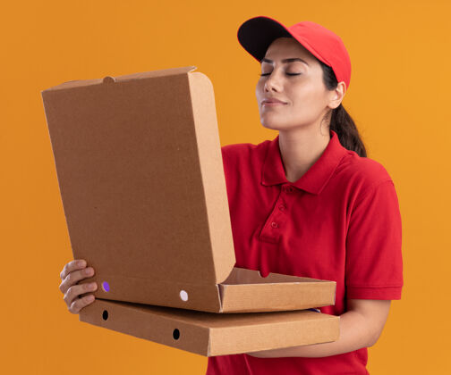 帽子年轻的送货女孩穿着制服 戴着帽子 嗅着隔离在橙色墙上的披萨盒穿年轻嗅