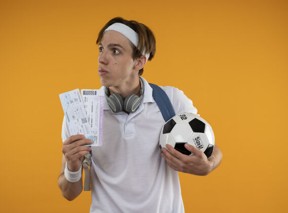 运动思想年轻的运动家伙戴着头带与腕带和背包与耳机在脖子上拿着票与球隔离在黄色墙上背包家伙票