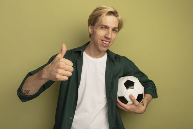 秀微笑着的金发小伙子穿着绿色t恤拿着球 竖起大拇指大拇指微笑抱着