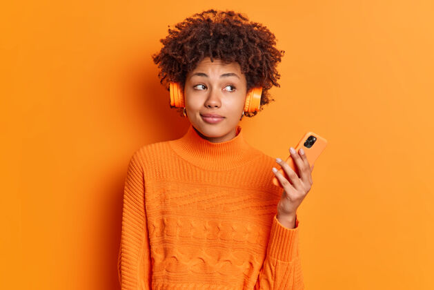 黑色体贴的女性梅洛曼听着播放列表中的音频曲目 抱着现代智能手机沉思地看着一旁满意微笑女人