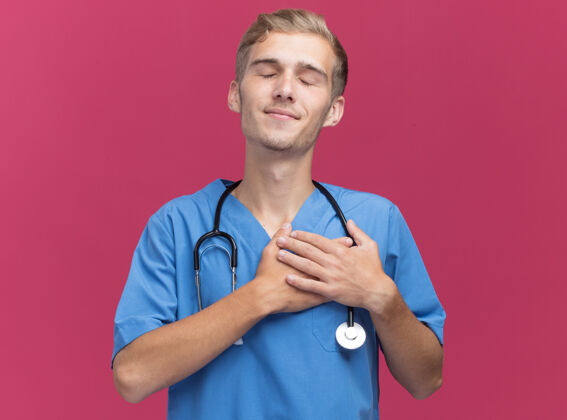 医生喜欢闭着眼睛的年轻男医生穿着医生制服用听诊器把手放在心上隔离在粉红色的墙上手封闭男性
