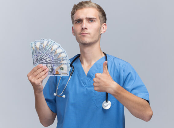 制服自信的年轻男医生穿着医生制服 手持听诊器拿着现金 在白墙上孤立地竖起大拇指现金显示信心