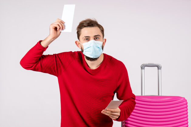 颜色正面图：戴着面具的年轻男性手持白墙上的飞机票病毒航行covid-航班旅行度假色病毒机票航班
