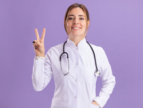 年轻面带微笑的年轻女医生穿着医用长袍 听诊器显示和平姿态 隔离在紫色的墙上表演和平女性
