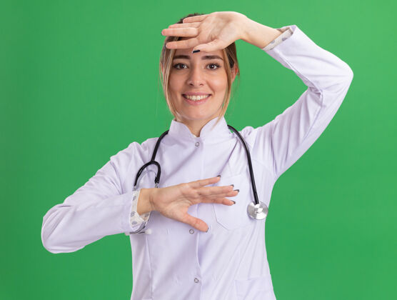 女性面带微笑的年轻女医生 穿着医用长袍 手举听诊器 额头被隔离在绿色的墙上抱着长袍手