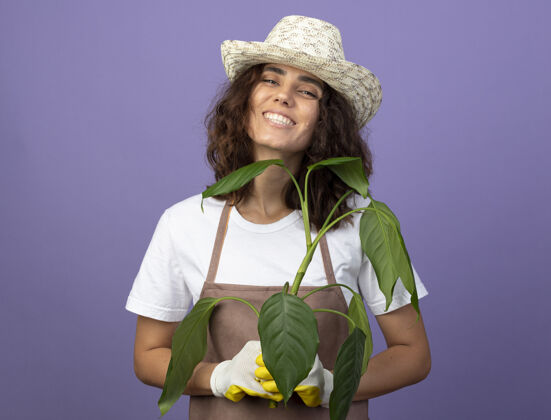 成人年轻的女园丁面带微笑 穿着制服 戴着园艺帽和手套 手里拿着植物园艺年轻制服
