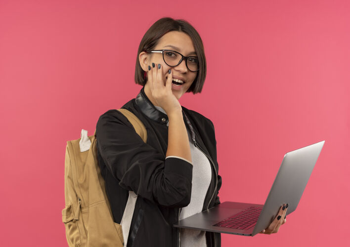 高兴快乐的年轻女学生戴着眼镜 背着书包拿着笔记本电脑 手放在脸颊上 隔离在粉色上抱背眼镜