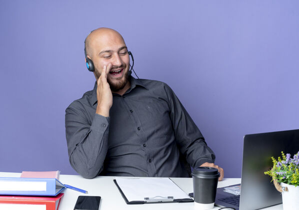坐着快乐的年轻秃头呼叫中心男子戴着耳机坐在办公桌旁 拿着工作工具看着笔记本电脑 把手放在离嘴不远的紫色上高兴近紫色