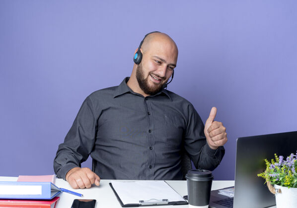 耳机快乐的年轻秃头呼叫中心男子戴着耳机坐在办公桌旁 拿着工作工具看着笔记本电脑 在紫色的地板上孤立地竖起大拇指紫色秃头工具