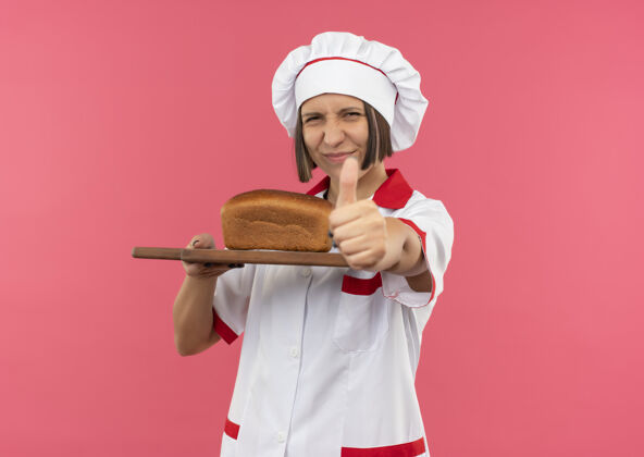 请身着厨师制服的年轻女厨师手拿着插有面包的砧板 在粉色背景上孤立地竖起大拇指 并留有复印空间制服拿着切着