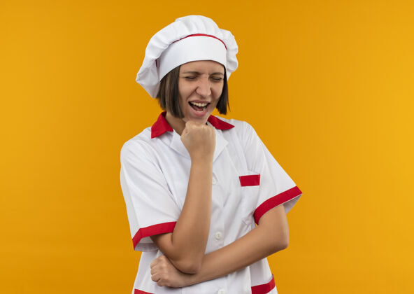 拳头快乐的年轻女厨师穿着厨师制服 把拳头放在下巴下 闭上眼睛孤立在橙色上橙色下巴封闭