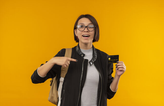 持有印象深刻的年轻女学生戴着眼镜 背着书包拿着信用卡指着橙色隔离印象佩戴眼镜
