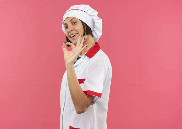 女身着厨师制服的快乐年轻女厨师站在侧视图中做ok标志孤立在粉色上制服烹饪立场