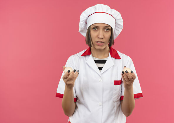 拿着穿着厨师制服的年轻女厨师拿着鸡蛋看着隔离在粉色背景上的相机 还有复制空间烹饪厨师年轻