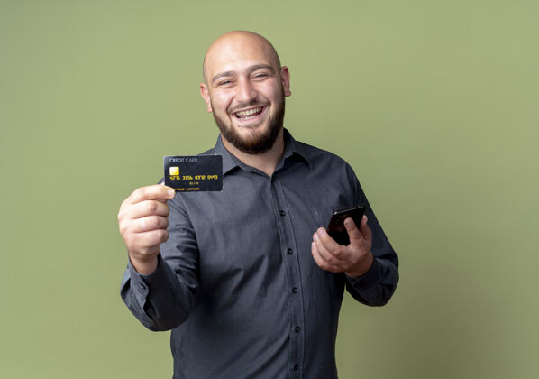男人快乐的年轻秃头呼叫中心男子手持手机 伸出信用卡孤立在橄榄绿手机信用喜悦