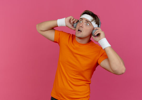 壁板印象深刻的年轻英俊的运动男子戴着头带和腕带和耳机手持耳机 看着一边孤立的粉红色腕带耳机头带