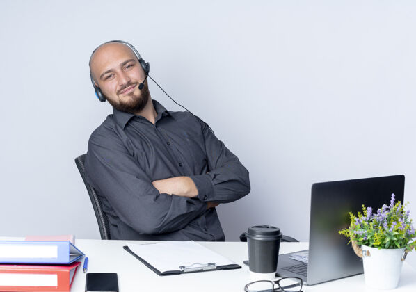 中心高兴的年轻秃头呼叫中心男子戴着耳机坐在办公桌与白色背景上孤立的工作工具封闭的姿势姿势耳机工作