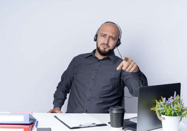 磨损戴着耳机的秃头呼叫中心年轻男子坐在办公桌旁 拿着工作工具 指着隔离在白色背景上的摄像机坐着年轻电话
