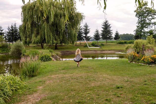 服装快乐的女孩儿站在美丽花园的池塘前美丽白天草