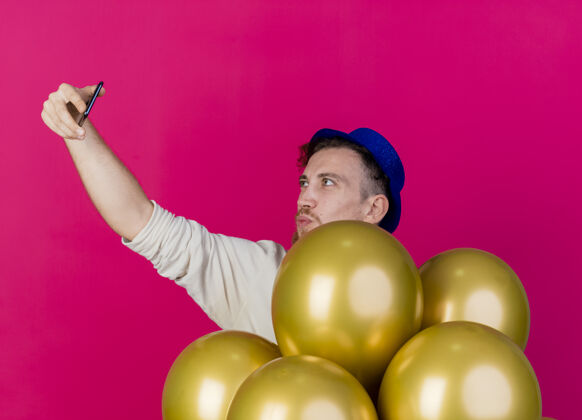 站着年轻英俊的斯拉夫党人戴着党的帽子站在气球后面做亲吻手势自拍隔离在粉红色的墙上亲吻拿着年轻