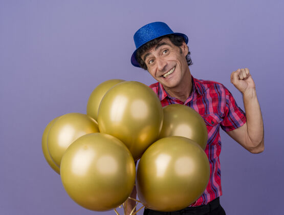 人微笑的中年派对男子戴着派对帽拿着气球看着边做是的手势孤立在紫色的墙上气球表情手势
