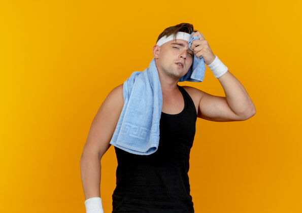 毛巾疲惫的年轻帅气的运动型男人戴着头带和腕带 脖子上围着毛巾 用隔离在橙色背景上的毛巾擦着汗水穿汗水周围