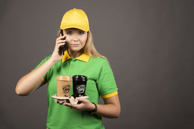 快递年轻的女送货员拿着几杯咖啡在智能手机上说高质量的照片杯子人制服
