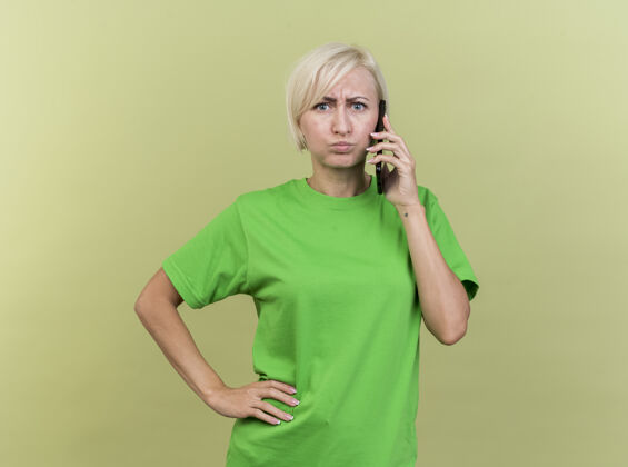 站皱眉的中年金发斯拉夫女人在讲电话 看着前面 双手放在腰上 隔离在橄榄绿的墙上 留着复印空间皱眉橄榄表情