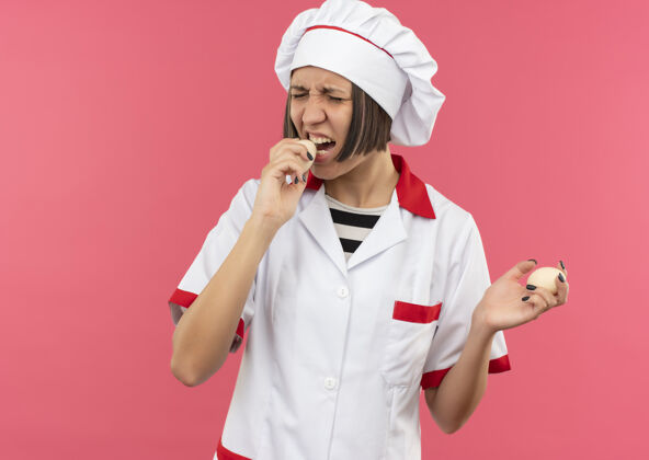 试试年轻的女厨师穿着厨师制服 拿着和试图咬鸡蛋隔离在粉红色背景与复制空间鸡蛋女咬
