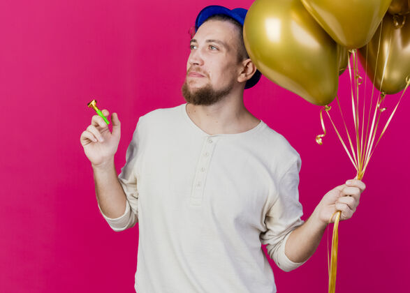 黄金年轻英俊的斯拉夫党人戴着党的帽子拿着气球和党吹在粉红色的墙上孤立一边看壁板气球公民