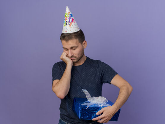 盒子戴着生日帽的悲伤帅哥把手放在下巴上 把礼盒孤立地放在紫色的墙上 留着复制空间礼物人生日