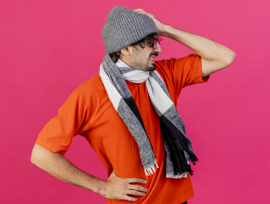 围巾戴着眼镜 戴着冬天的帽子 戴着围巾 手放在腰上 头放在头上 眼睛闭着 看着侧面 隔离在粉红色的墙上壁板姿势人