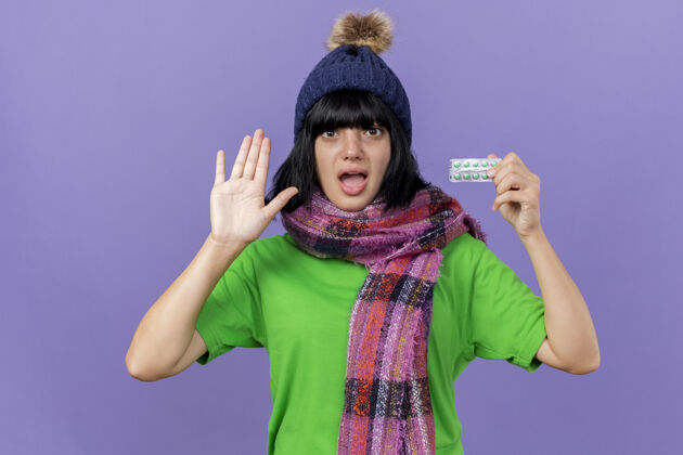 冬天印象深刻的年轻生病的女人戴着冬天的帽子和围巾看着前面拿着一包胶囊显示空手孤立在紫色的墙上表演包穿着