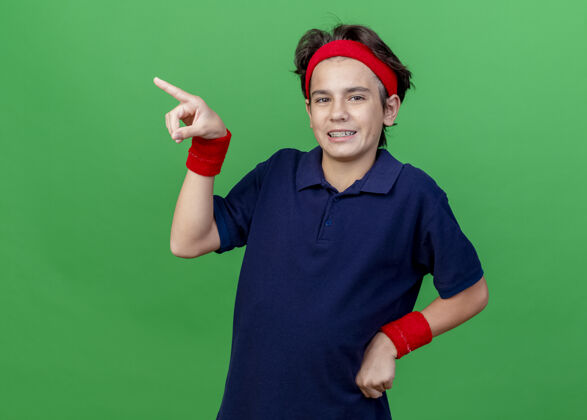 人面带微笑的年轻帅气的运动男孩戴着头带和带牙套的腕带 手放在腰上 看着前面 指着隔离在绿色墙壁上的一边 留着复制空间运动感觉腰