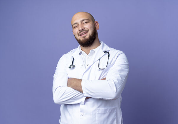 微笑微笑的年轻秃头男医生穿着医用长袍和听诊器交叉双手隔离在蓝色背景上长袍男性听诊器