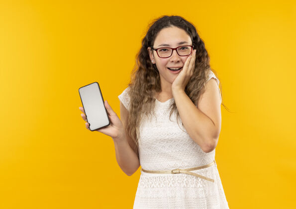 女生印象深刻的年轻漂亮的女学生戴着眼镜背着包拿着手机把手放在脸颊上隔离在黄色抱年轻印象