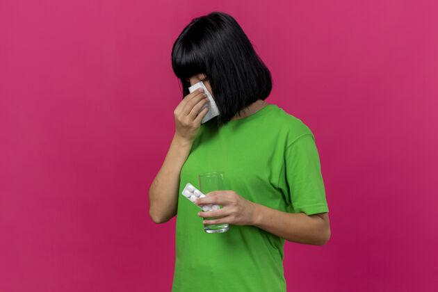 疾病年轻的患病妇女站在侧视图中 拿着药片和一杯水 用餐巾擦鼻子 隔离在粉红色的墙上 留有复印空间湿巾水脸
