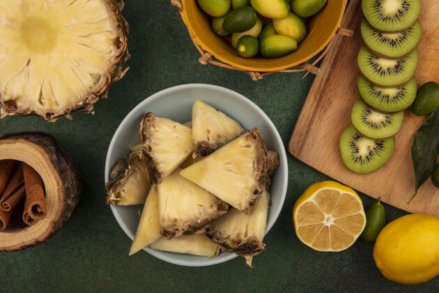 棒顶视图甜菠萝片放在碗上 猕猴桃片放在木制厨房板上 金盏花放在桶上 肉桂棒和柠檬隔离在绿色背景上木材新鲜特写