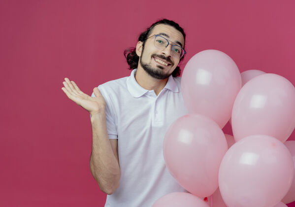 帅气笑容可掬的帅哥戴着眼镜拿着气球 用手指着隔离在粉色背景上的复制空间微笑穿拿着