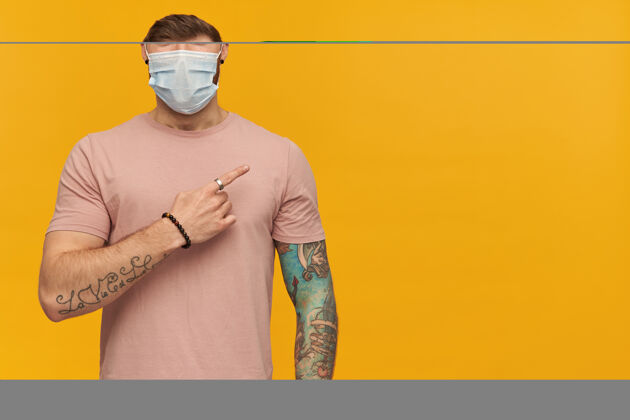 学生戴着防冠状病毒的粉色T恤和防病毒面具的快乐的纹身年轻人 用手指指着黄色墙壁上的侧边男人空虚健康
