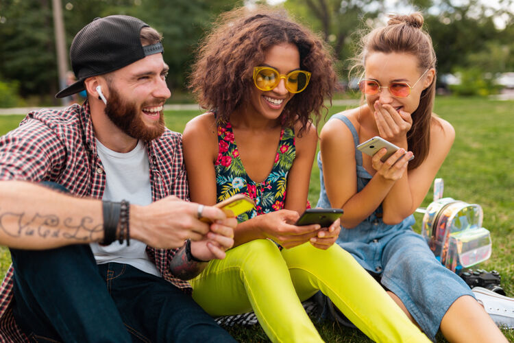 眼镜快乐的年轻朋友们坐在公园里用智能手机微笑 男人和女人一起玩耳机兴奋时尚