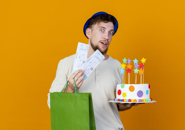 生日令人印象深刻的年轻英俊的斯拉夫党家伙戴着党的帽子拿着机票纸袋和生日蛋糕与明星看着相机隔离在橙色背景与复制空间票斯拉夫包